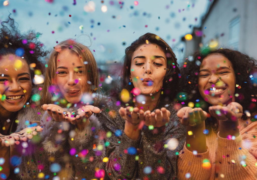 Young women blowing confetti from hands. Young women blowing confetti from hands. Friends celebrating outdoors in evening at a terrace.